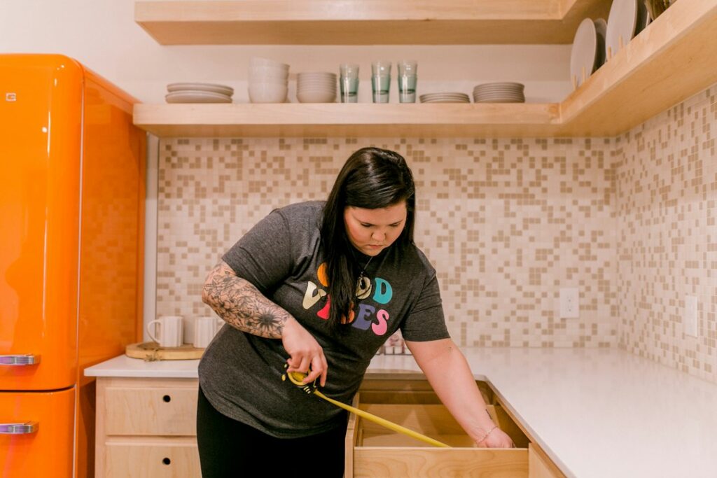Homeowner measures kitchen cabinets while reviewing storage, lighting, appliances, and walkway space.