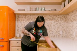 Homeowner measures kitchen cabinets while reviewing storage, lighting, appliances, and walkway space.